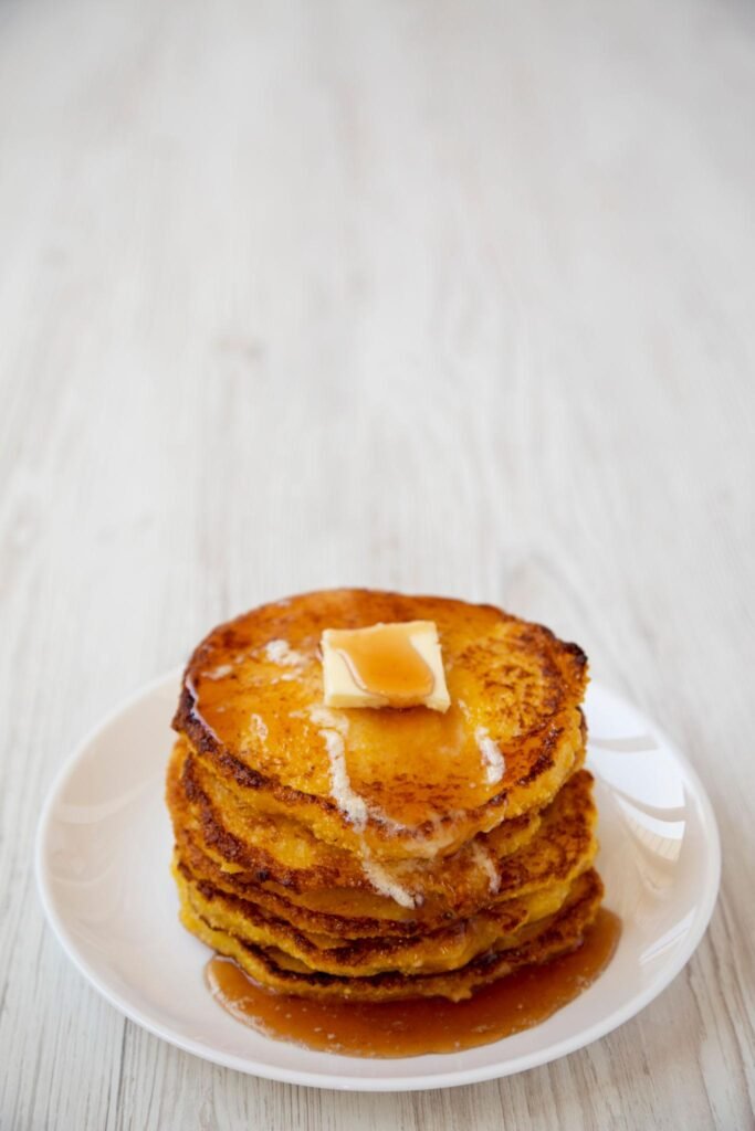 Four golden-brown cornmeal johnnycakes stacked on a ceramic plate with melting butter and maple syrup.